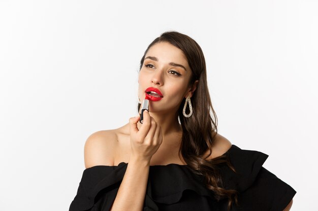 Concepto de moda y belleza. Hermosa mujer vestida de negro aplicando lápiz labial rojo y maquillaje, yendo de fiesta, de pie sobre fondo blanco.