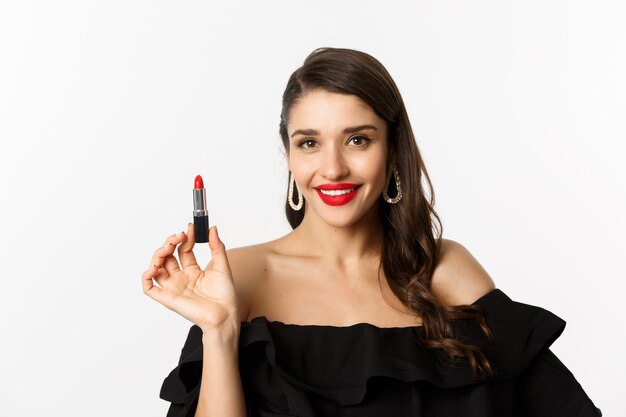 Concepto de moda y belleza. Hermosa mujer en vestido negro aplicando lápiz labial rojo y maquillaje, yendo de fiesta, de pie sobre fondo blanco.