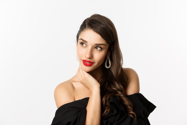 Concepto de moda y belleza. Mujer elegante con aretes y vestido negro, mirando a la izquierda sensual, de pie sobre fondo blanco.