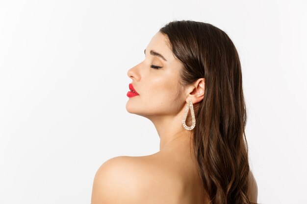 Concepto de moda y belleza. Perfil de hermosa mujer desnuda de pie con maquillaje y aretes, mirando tierna y sensual, de pie sobre fondo blanco.