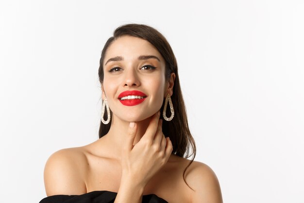 Concepto de moda y belleza. Primer plano de una hermosa mujer morena con labios rojos, tocando la cara y sonriendo seguro de sí mismo, de pie sobre fondo blanco.