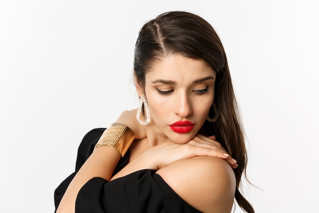 Concepto de moda y belleza. Primer plano de mujer sensual en elegantes aretes y vestido negro, con maquillaje con labios rojos, mirando hacia abajo con ternura, de pie sobre fondo blanco.