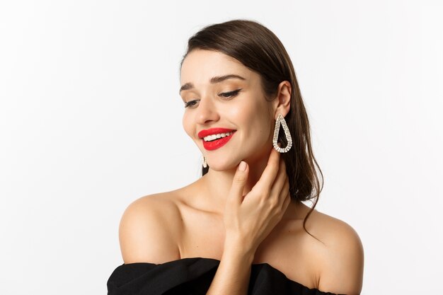 Concepto de moda y belleza. Primer plano de mujer tierna en vestido negro y aretes, tocando suavemente la cara y sonriendo, mirando hacia abajo coqueta, de pie sobre fondo blanco.