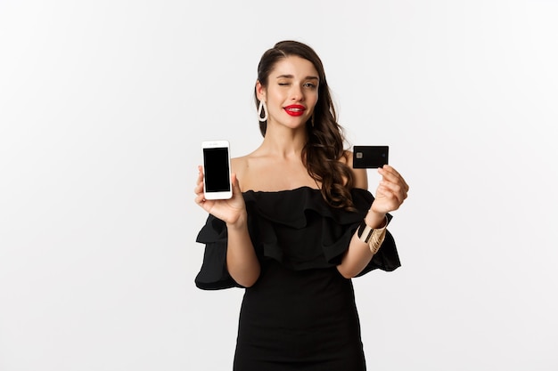 Concepto de moda y compras. Hermosa mujer con labios rojos, guiñando un ojo a la cámara, mostrando la pantalla del teléfono inteligente y la tarjeta de crédito, comprando en línea, fondo blanco.