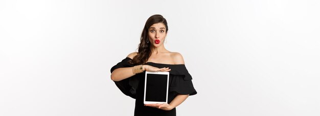 Concepto de moda y compras hermosa mujer con lápiz labial rojo vestido negro mostrando una pantalla de tableta