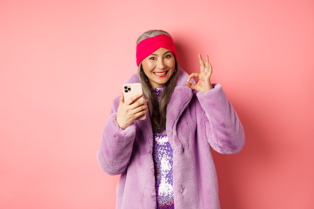 Concepto de moda y compras en línea. Mujer mayor asiática elegante que muestra el signo correcto y que sostiene el teléfono móvil, recomendando la tienda de Internet, fondo rosado