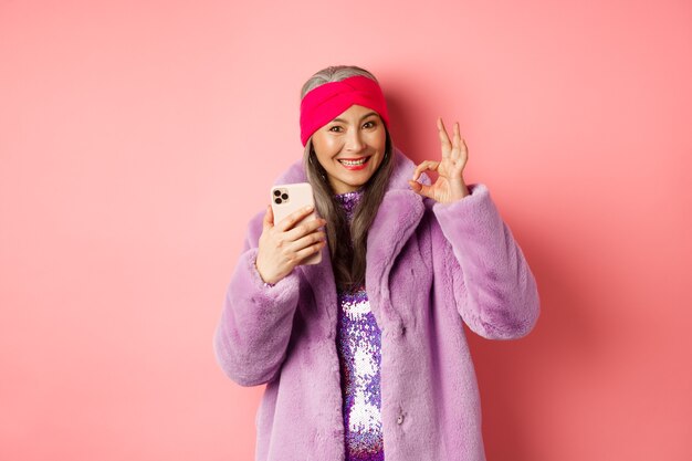 Concepto de moda y compras en línea. Mujer mayor asiática elegante que muestra el signo correcto y que sostiene el teléfono móvil, recomendando la tienda de Internet, fondo rosado