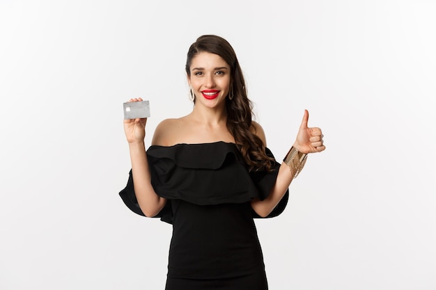 Concepto de moda y compras. Mujer elegante satisfecha en vestido negro mostrando tarjeta de crédito, hacer pulgar hacia arriba en señal de aprobación, recomendando, de pie sobre fondo blanco.