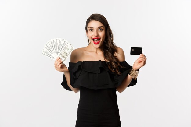 Concepto de moda y compras. Mujer emocionada en vestido negro, mostrando tarjeta de crédito y dólares, sonriendo y mirando a cámara, fondo blanco.