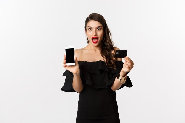 Concepto de moda y compras. Mujer hermosa asombrada que muestra la pantalla del teléfono inteligente y la tarjeta de crédito, mirando emocionado, comprando en línea, fondo blanco.