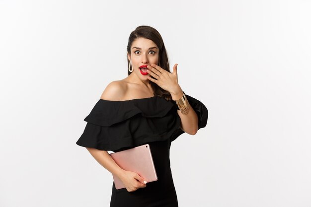 Concepto de moda y compras. Mujer joven elegante con maquillaje glamour, vestido negro, sosteniendo tableta digital y mirando sorprendido, fondo blanco.