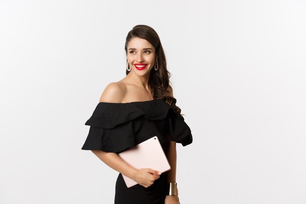 Concepto de moda y compras. Mujer joven con estilo con maquillaje glamour, vestido negro, sosteniendo tableta digital y sonriendo, fondo blanco.