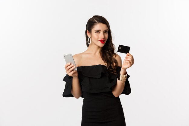 Concepto de moda y compras. mujer con labios rojos, vestido negro, pensando qué comprar, sosteniendo una tarjeta de crédito y un teléfono móvil, de pie sobre un fondo blanco.