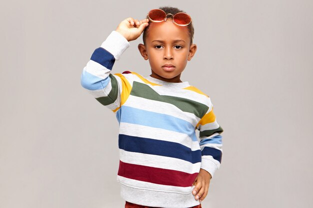 Concepto de moda, estilo, ropa infantil y accesorios para niños. Grave confianza muchacho afroamericano modelado contra la pared en blanco vistiendo jersey de rayas y tonos rosados en la cabeza