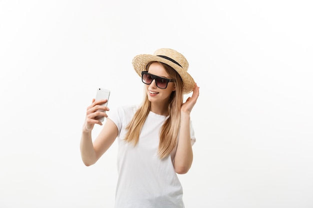Concepto de moda y estilo de vida: mujer joven y bonita con sombrero, gafas de sol tomando una foto de sí misma por teléfono móvil aislado sobre fondo blanco.