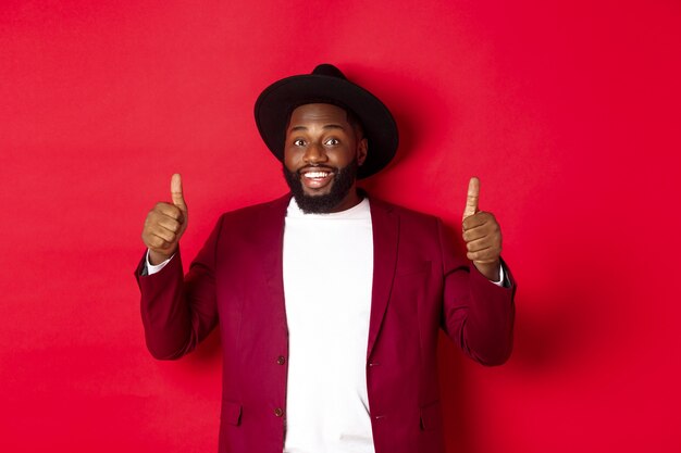 Concepto de moda y fiesta. Alegre hombre afroamericano en elegante chaqueta, celebrando el año nuevo de Navidad, mostrando los pulgares hacia arriba y sonriendo, como y aprobar, fondo rojo.