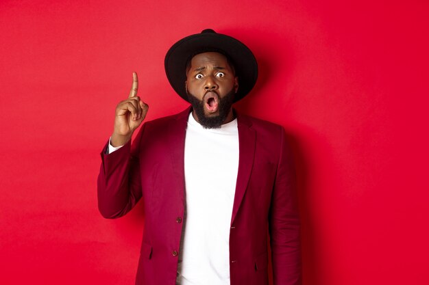 Concepto de moda y fiesta. Hombre negro emocionado que tiene una idea, levantando el dedo para decir sugerencia, de pie con elegante sombrero y chaqueta, fondo rojo.