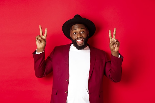 Concepto de moda y fiesta. hombre negro guapo divirtiéndose, mostrando signos de paz y sonriendo, de pie con sombrero sobre fondo rojo.
