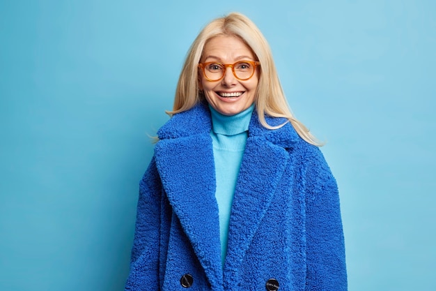 Concepto de moda de invierno. Feliz sonriente mujer rubia de mediana edad lleva gafas ópticas y abrigo azul.