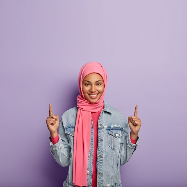 Concepto de moda islámica. Alegre mujer positiva con apariencia y ropa específicas, puntos arriba en el espacio libre, muestra algo hacia arriba, usa una chaqueta fashionabe. Chica con hijab anuncia objeto