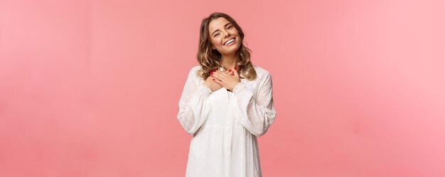 Concepto de moda y mujer de belleza Retrato de mujer joven rubia femenina en vestido blanco lindo sostener los brazos presionados al corazón y suspirando de admiración y deleite sentirse agradecido fondo rosa