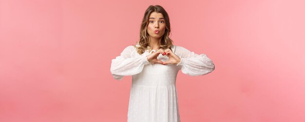 Concepto de moda y mujeres de belleza Retrato de una tierna y encantadora jovencita rubia con un hermoso vestido blanco que dobla los labios en un beso y hace un signo de corazón cerca del pecho de pie con fondo rosa muestra simpatía