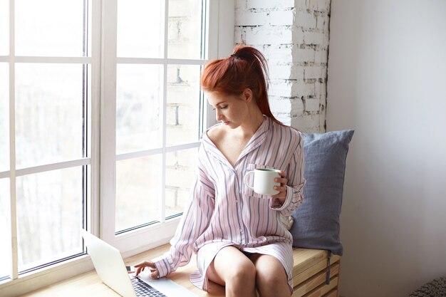 Concepto moderno de tecnología, ocio y comunicación. Chica bonita pelirroja con cola de caballo sentada en el alféizar de la ventana con una taza de bebida caliente, utilizando una conexión a Internet de alta velocidad en una computadora portátil genérica