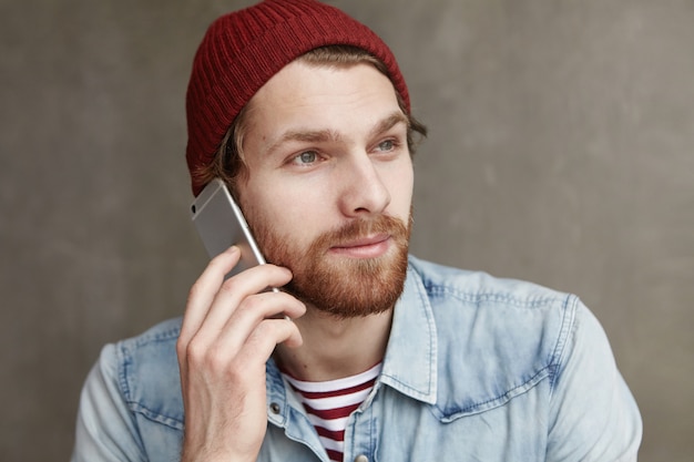 Concepto moderno de tecnología, personas y comunicación. Disparo en la cabeza del estudiante barbudo de moda con sombrero y camisa vaquera que tiene una conversación agradable en el teléfono inteligente, sonriendo mientras recibe noticias positivas