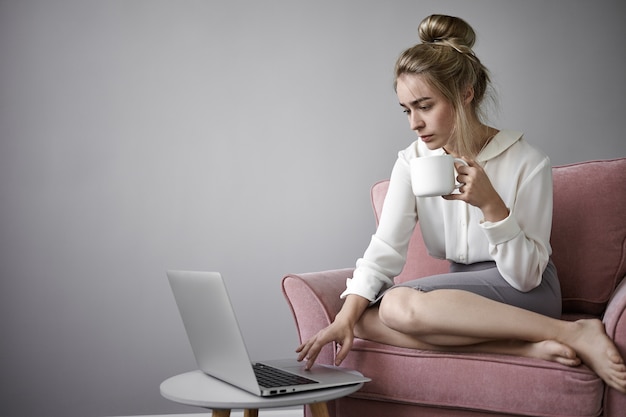 Concepto moderno de tecnología, trabajo y ocio. Grave joven empresaria tomando café por la mañana y revisando el correo electrónico antes de ir a la oficina, sentada en un sillón frente a una computadora portátil abierta, sosteniendo una taza grande