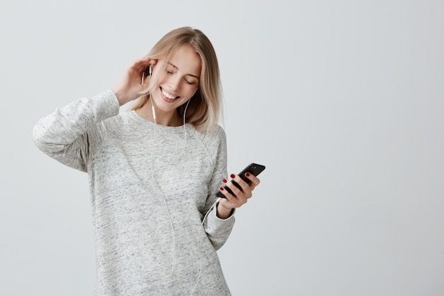 Concepto de música, felicidad y tecnología. La encantadora mujer adolescente tiene el cabello teñido de rubio, escucha música en el teléfono celular y baila alegremente