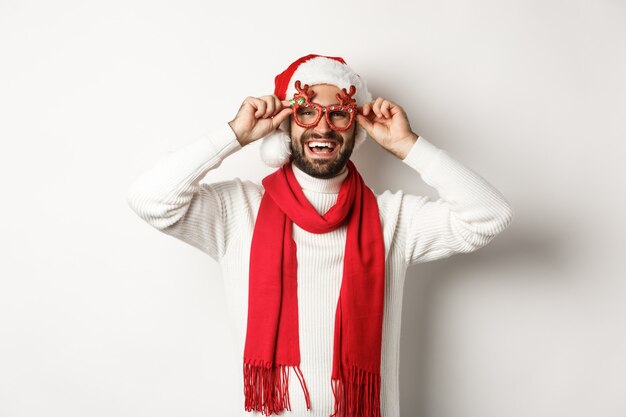 Concepto de Navidad, año nuevo y celebración. Hombre feliz riendo, vistiendo gorro de Papá Noel y gafas de fiesta, de pie sobre fondo blanco.