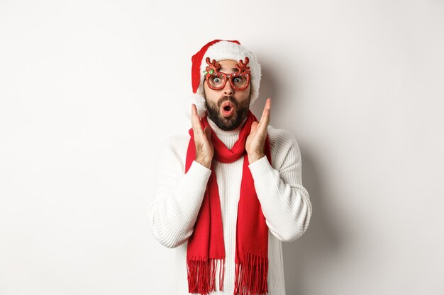 Concepto de Navidad, año nuevo y celebración. Hombre con gafas de fiesta y gorro de Papá Noel mirando sorprendido, escuchar oferta comercial, de pie sobre fondo blanco.