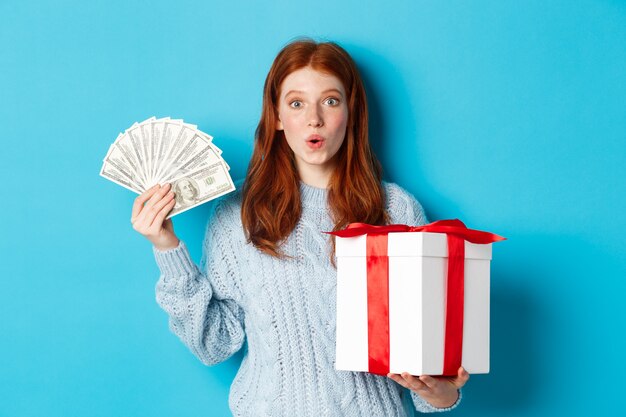 Concepto de Navidad y compras. Chica pelirroja emocionada mirando a cámara, sosteniendo un gran regalo de año nuevo y dólares, comprando regalos, de pie sobre fondo azul.