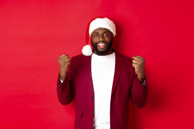 Concepto de Navidad, fiesta y vacaciones. Hombre negro emocionado celebrando el año nuevo, con gorro de Papá Noel, apretando los puños satisfechos, ganando y regocijándose, de pie contra el fondo rojo.