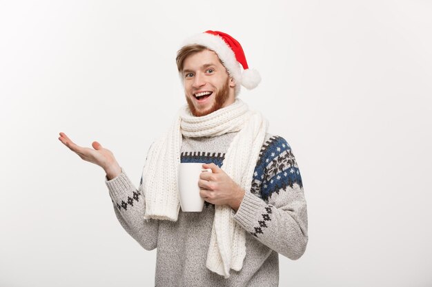 Concepto de Navidad Hombre joven con barba en suéter y gorro de Papá Noel sosteniendo una taza de café caliente apuntando con la mano al lado aislado en blanco con espacio de copia