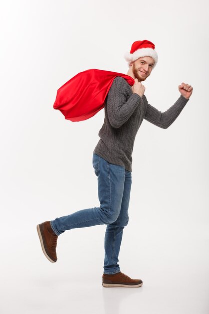 Concepto de navidad Joven hombre de barba confiado corriendo y sosteniendo una gran bolsa roja de santa con un montón de regalos dentro