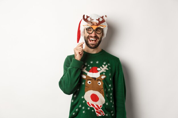 Concepto de Navidad y vacaciones. Hombre de barba sonriente con sombrero de Santa mirando feliz, sosteniendo la máscara de fiesta para el año nuevo, celebrando la Navidad, de pie sobre fondo blanco.