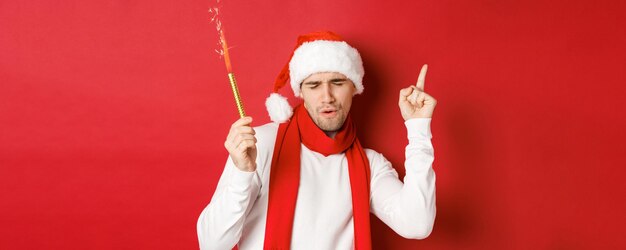 Concepto de Navidad, vacaciones de invierno y celebración. Chico atractivo disfrutando de la fiesta de año nuevo, bailando con bengala, vistiendo gorro de Papá Noel y bufanda, de pie sobre fondo rojo.