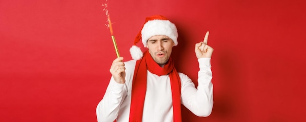 Concepto de Navidad, vacaciones de invierno y celebración. Chico atractivo disfrutando de la fiesta de año nuevo, bailando con bengala, vistiendo gorro de Papá Noel y bufanda, de pie sobre fondo rojo.