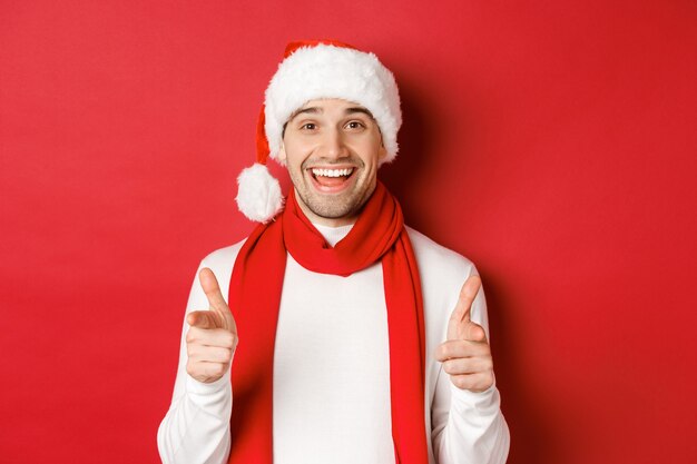 Concepto de Navidad, vacaciones de invierno y celebración. Hombre descarado con gorro de Papá Noel y bufanda, sonriendo y señalando con el dedo a la cámara, deseando feliz año nuevo, de pie sobre fondo rojo.