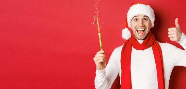 Concepto de navidad, vacaciones de invierno y celebración. Un hombre guapo celebrando el año nuevo y divirtiéndose, sosteniendo una bengala y mostrando el pulgar hacia arriba, de pie sobre un fondo rojo