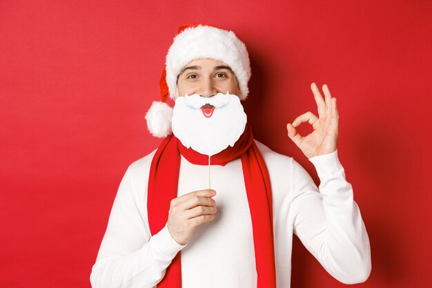 Concepto de Navidad, vacaciones de invierno y celebración. Hombre guapo complacido con gorro de Papá Noel, sosteniendo una larga máscara de barba blanca y mostrando un signo bien, de pie sobre fondo rojo