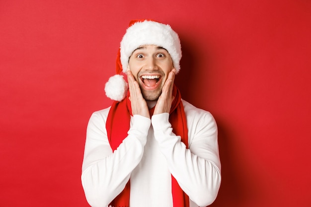 Concepto de Navidad, vacaciones de invierno y celebración. Primer plano de hombre sorprendido y feliz con gorro de Papá Noel y bufanda, mirando algo increíble, de pie sobre fondo rojo.