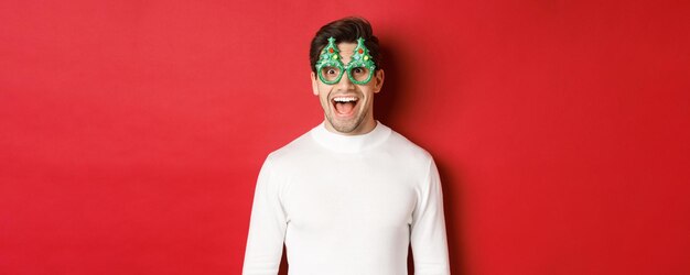 Concepto de Navidad, vacaciones de invierno y celebración. Primer plano de un joven apuesto disfrutando del año nuevo, con gafas de fiesta y mirando asombrado a la cámara, de pie sobre fondo rojo.