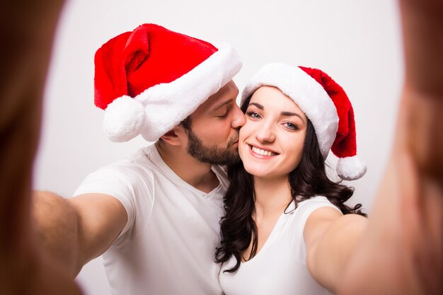 Concepto de Navidad, vacaciones, tecnología y personas - pareja feliz con sombreros de santa tomando foto selfie de manos sobre fondo blanco. Ellos miran la camara y se besan