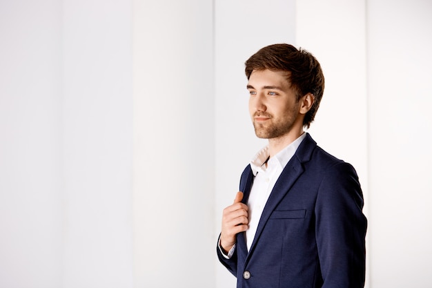 Concepto de negocio, éxito y personas. Apuesto hombre de negocios elegante en traje, soporte de oficina, disfrutando de la vista desde la ventana, sonriendo complacido