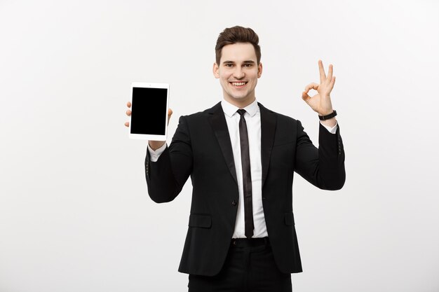 Concepto de negocio: hombre de negocios alegre en traje elegante con tablet pc mostrando ok. Aislado sobre fondo gris.