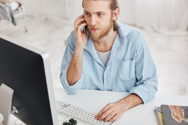 Concepto de negocio, oficina y tecnología. Vista superior del empleado barbudo con camisa azul, hablando por teléfono con compañeros, escribiendo en el teclado, mirando en la pantalla de la computadora, utilizando dispositivos modernos