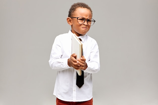 Concepto de niños, aprendizaje, educación y conocimiento. Retrato de niño africano enojado con camisa blanca, corbata y gafas, sosteniendo un cuaderno y haciendo muecas, enojado porque no sabe hacer matemáticas