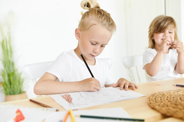 Concepto de niños, arte, creatividad y afición. Colegial rubio concentrado en camiseta blanca sosteniendo un lápiz negro, dibujando algo con diligencia, su hermana pequeña sonriendo sentada junto a él en el escritorio